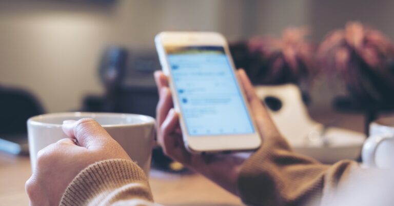 A person wearing a sweater sitting at a desk, holding a white mug in one hand and a phone in the other.