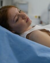 A young woman wearing a white tank top lying on a medical exam bed with a blue sheet in an examination room.
