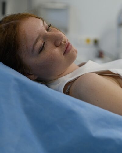 A young woman wearing a white tank top lying on a medical exam bed with a blue sheet in an examination room.