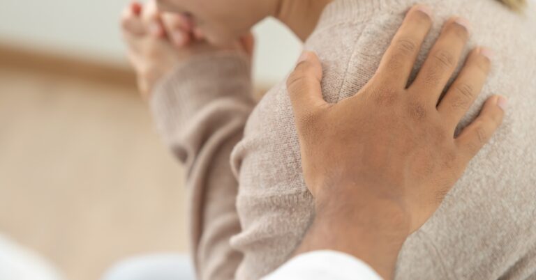 A woman sitting with her hands near her face in a nervous posture, while someone else's hand rests on her shoulder.