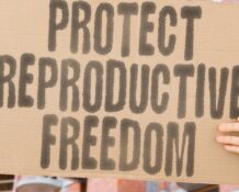 A person standing in front of a row of buildings, holding a cardboard sign with the message 