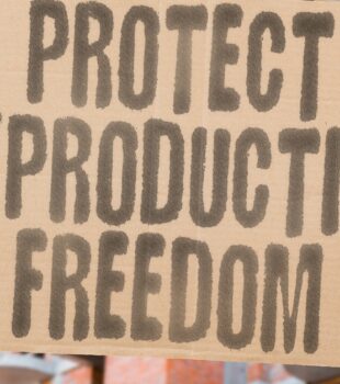 A person standing in front of a row of buildings, holding a cardboard sign with the message 