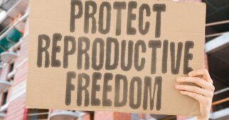 A person standing in front of a row of buildings, holding a cardboard sign with the message 