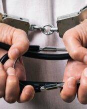 A man in a white lab coat shown from behind. His hands are in handcuffs, and he's holding a folded stethoscope.