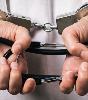 A man in a white lab coat shown from behind. His hands are in handcuffs, and he's holding a folded stethoscope.