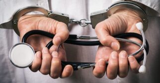 A man in a white lab coat shown from behind. His hands are in handcuffs, and he's holding a folded stethoscope.