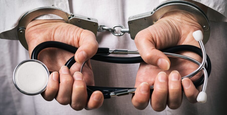 A man in a white lab coat shown from behind. His hands are in handcuffs, and he's holding a folded stethoscope.