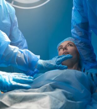A woman dressed in a gown and hair net lying on a surgical table, surrounded by several other people in full scrubs.