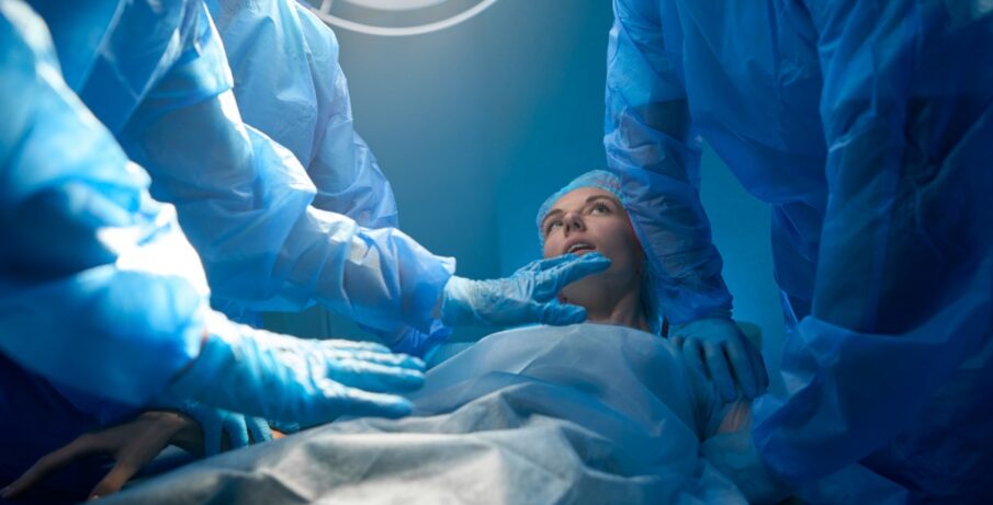 A woman dressed in a gown and hair net lying on a surgical table, surrounded by several other people in full scrubs.