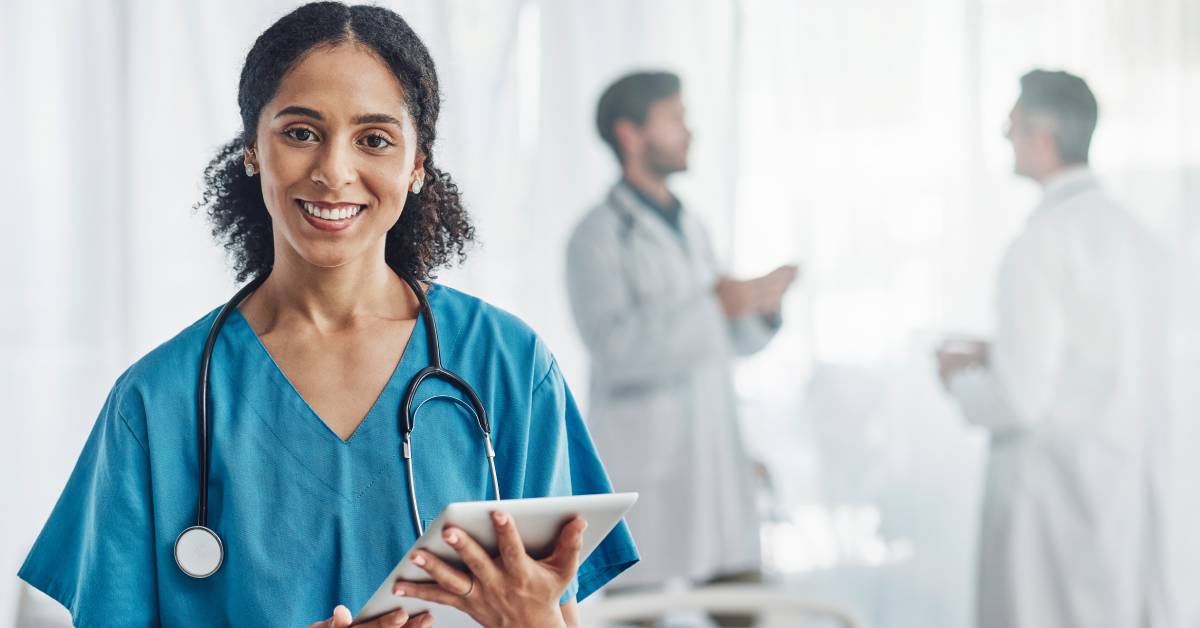 A woman healthcare worker holding a tablet and smiling while wearing a stethoscope in a hospital setting.