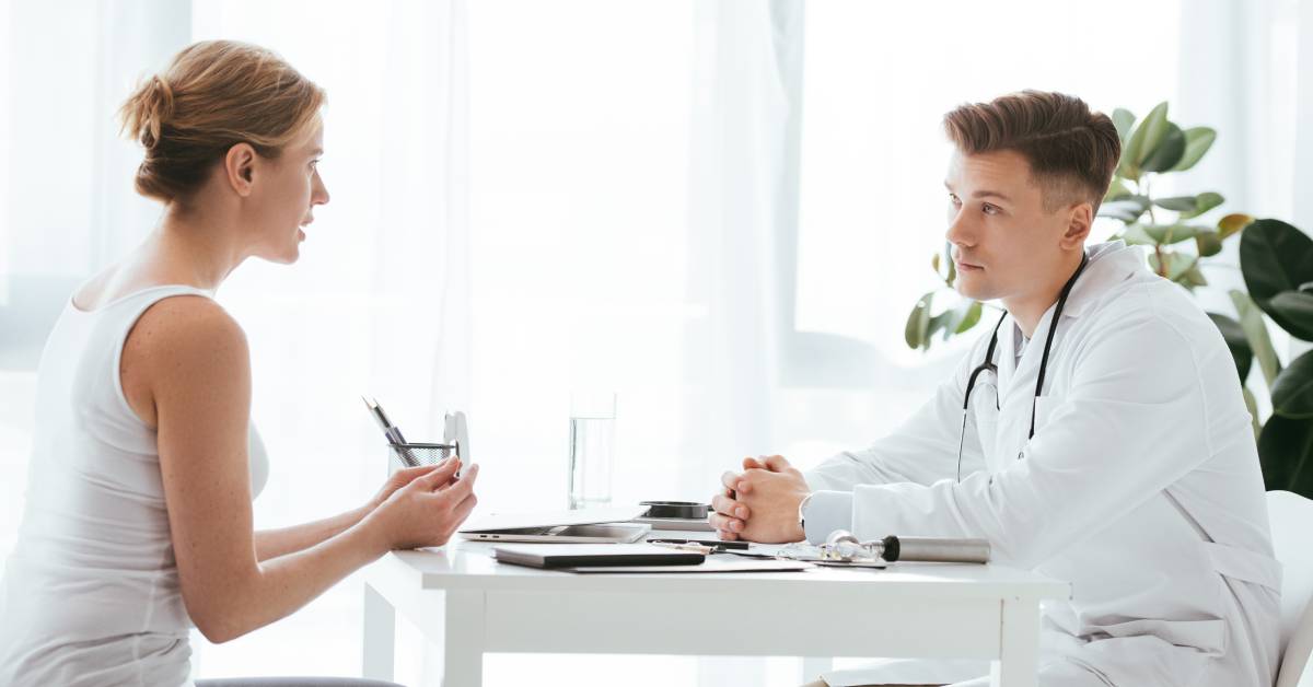 A doctor in a white coat sitting at a desk and talking with a patient seated across from them in a clinic room.