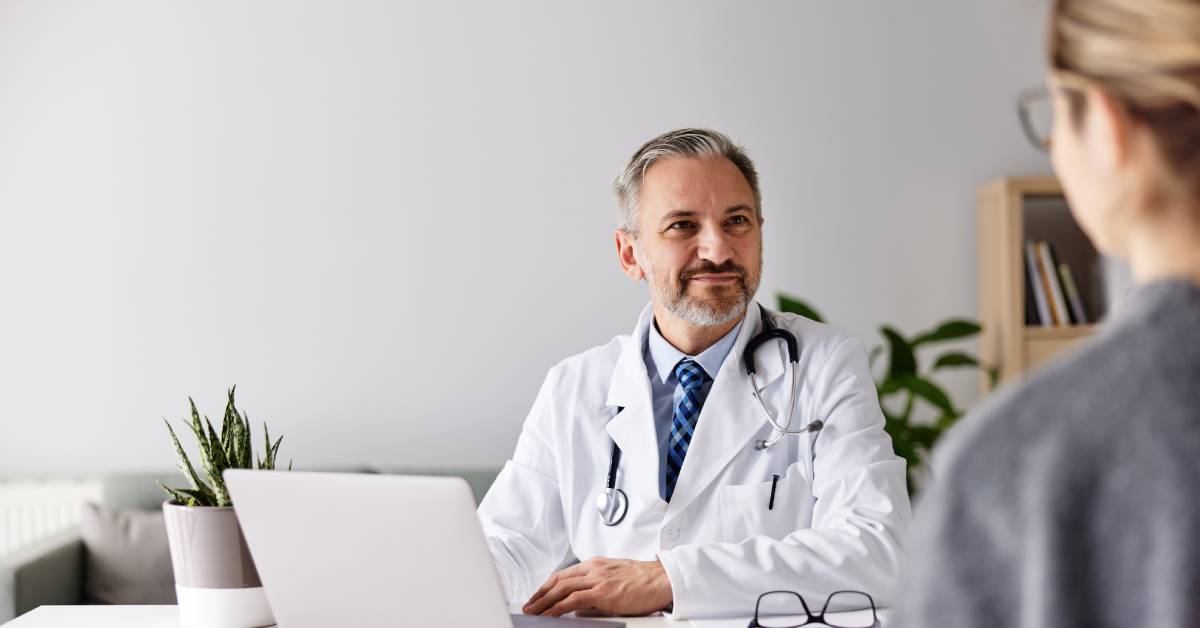 A male doctor sitting and talking with a female patient across a desk inside a medical office setting.