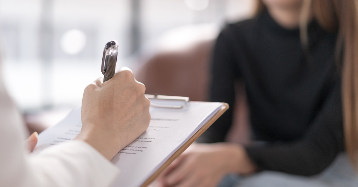 A woman sitting across from a person in a white lab coat holding a clipboard with paper and taking notes.