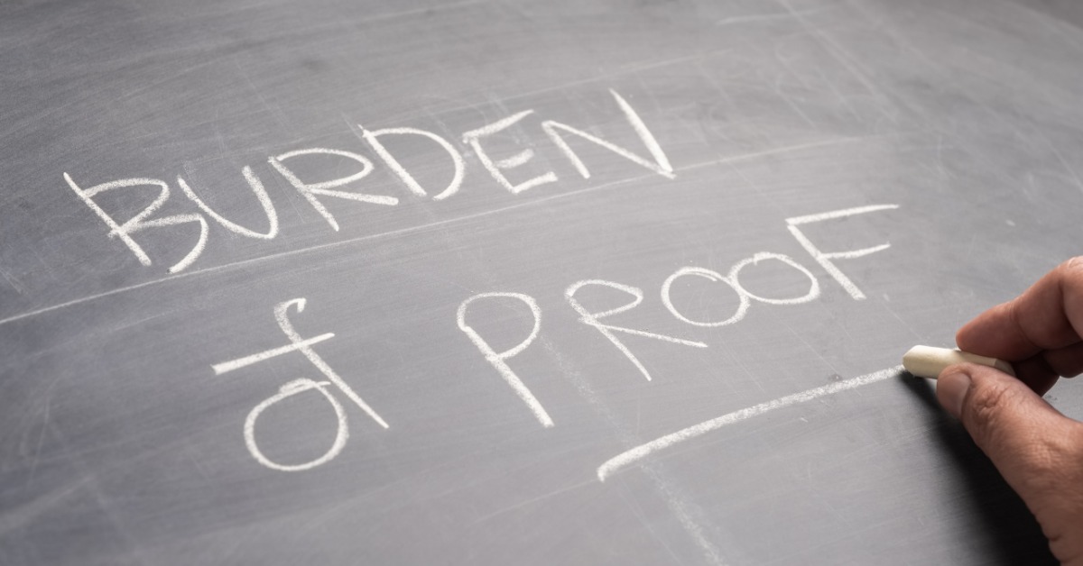 A hand holding white chalk near the words Burden of Proof written on a dark chalkboard surface in close view.