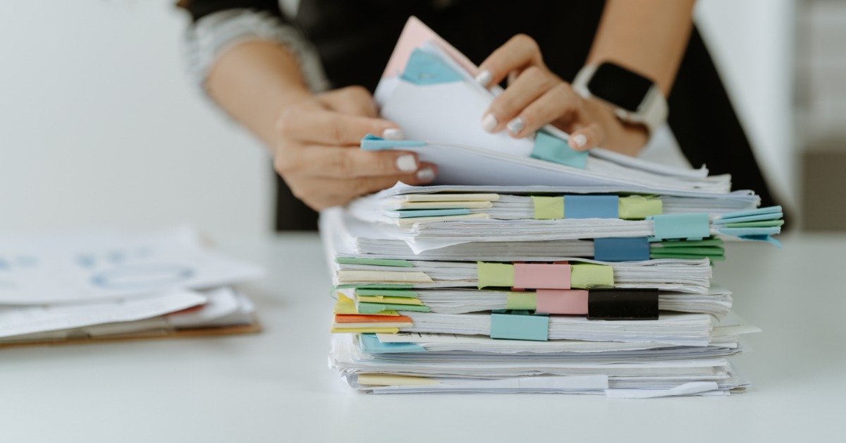 A person sorting through stacks of paper forms and folders spread across a desk during office work at a table.