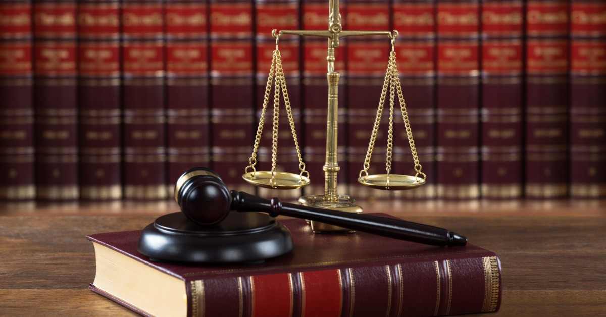 A wooden mallet resting beside a large, bulky legal book and a brass justice scale on a table in a law office.