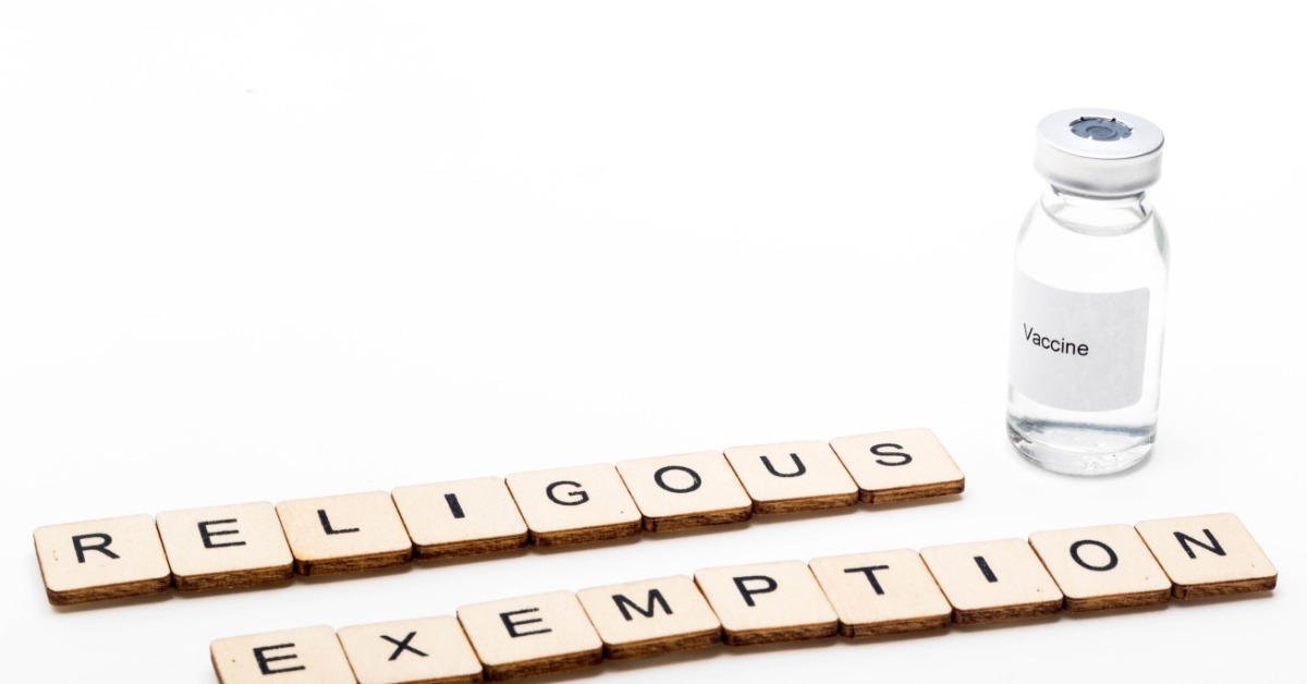 A glass vial with a label reading "vaccine" and letter blocks spelling "religious exemption" on a plain white surface.