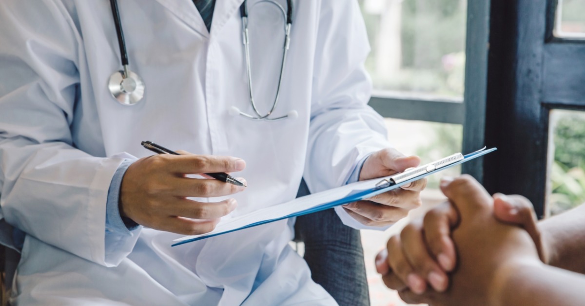 A person in a white coat explaining paperwork to another person seated across a desk in a medical office.