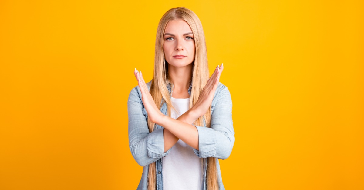 A blond woman standing with crossed arms and a serious expression against a bright yellow background.