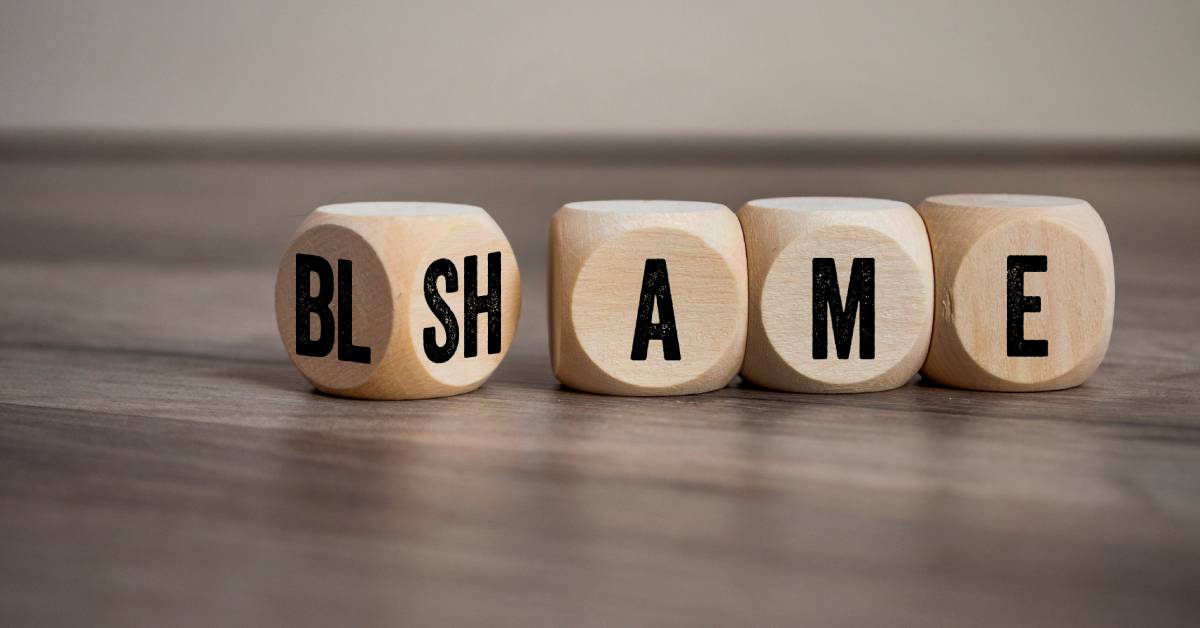 Wooden cubes on a wooden surface showing black letters that spell blame/shame against a blurred neutral background.