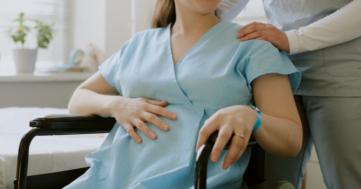 A pregnant woman in a blue gown sitting in a wheelchair while another person rests a hand on her shoulder.