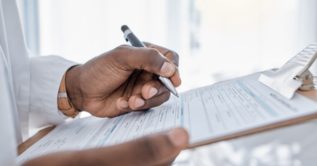 A hand holding a pen over a medical form on a clipboard, with a white coat sleeve and bright office background.