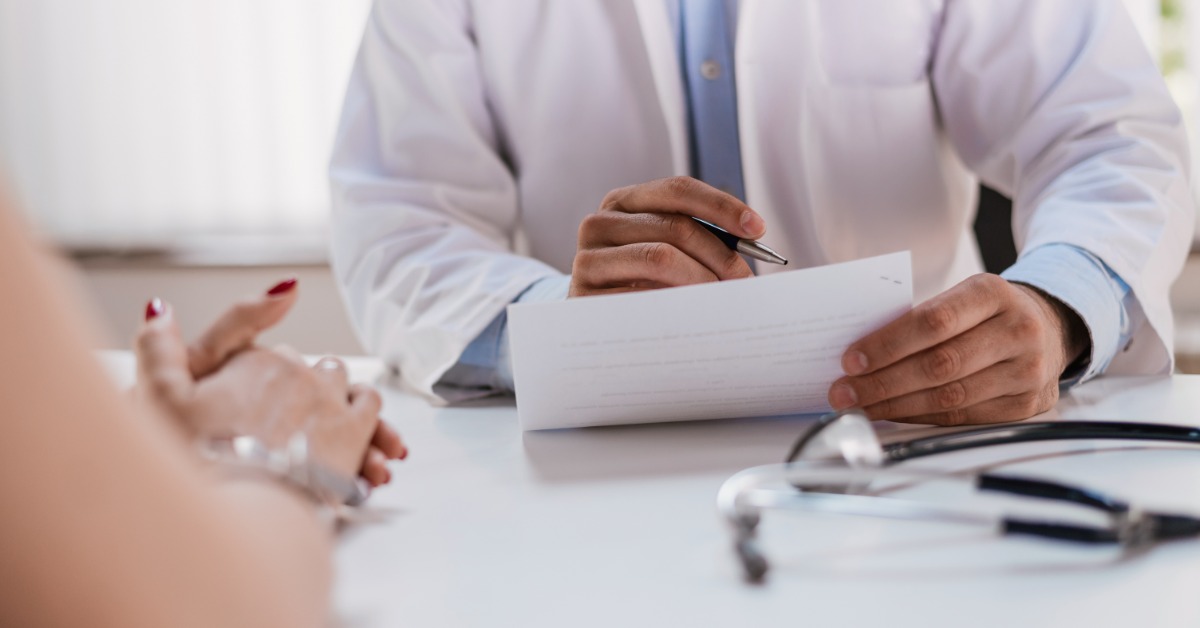 A person in a white coat holding a pen and paper across from another person at a desk with a stethoscope.