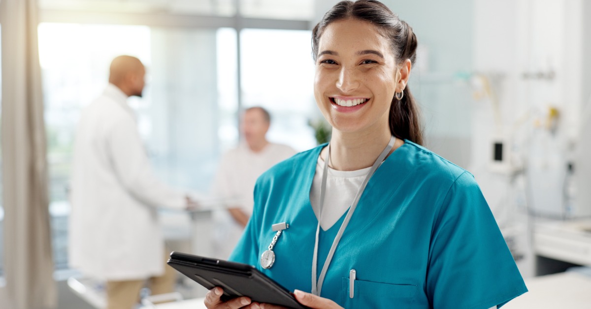 A woman in teal scrubs smiling while holding a tablet in a bright medical office with two people behind her.