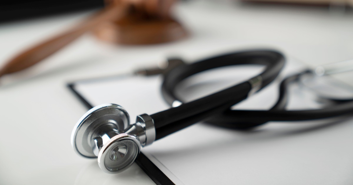 A stethoscope resting on a document with a wooden gavel blurred in the background on a white table surface.
