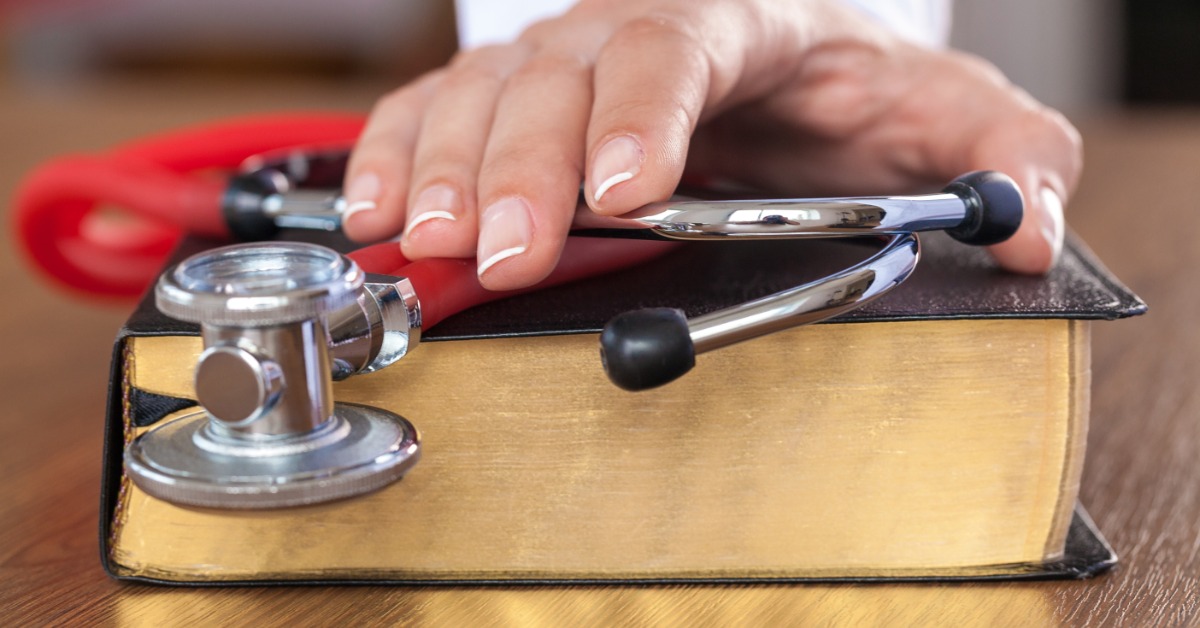 A hand resting on a stethoscope placed across a thick book on a wooden table, with red tubing nearby.