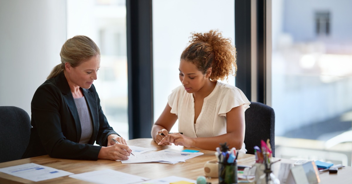 Two women sitting at a wooden office table, writing on documents with papers and colorful notes spread around.