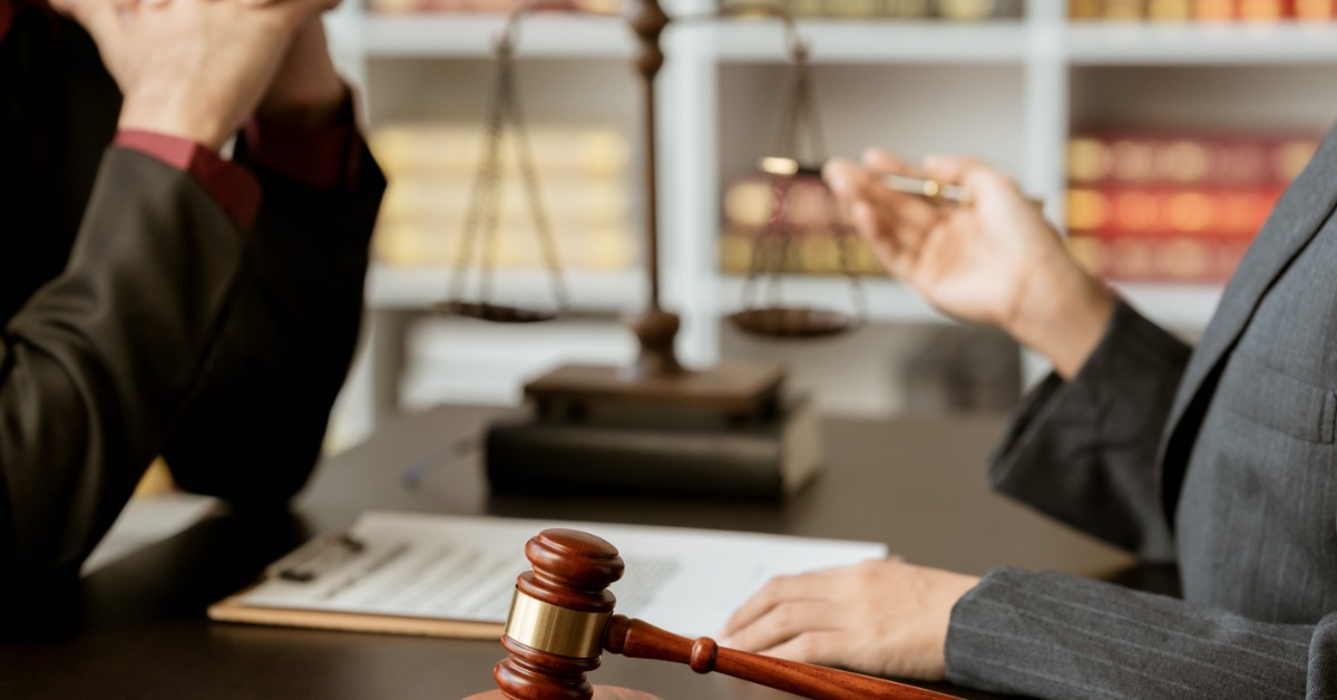Two people sitting across a desk with a wooden gavel, documents, scales, and books visible between them.