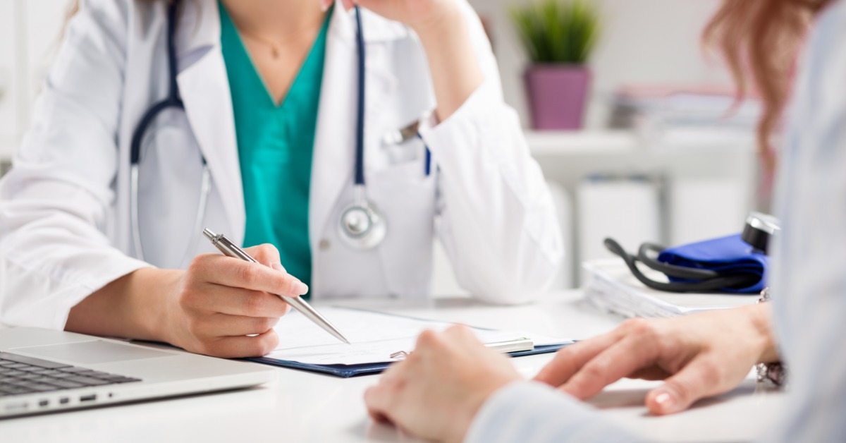 A person in a white coat holding a pen beside documents while another person sits across a desk in an office.