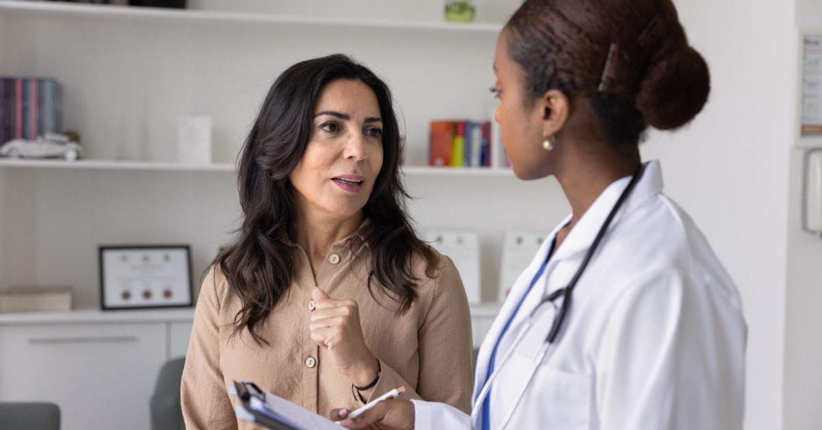  A woman speaking with a person in a white coat holding a clipboard inside a bright medical office with shelves.