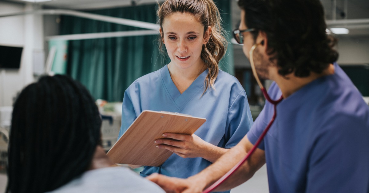  A person in blue scrubs holding a clipboard while another uses a stethoscope near a seated patient in a medical room.