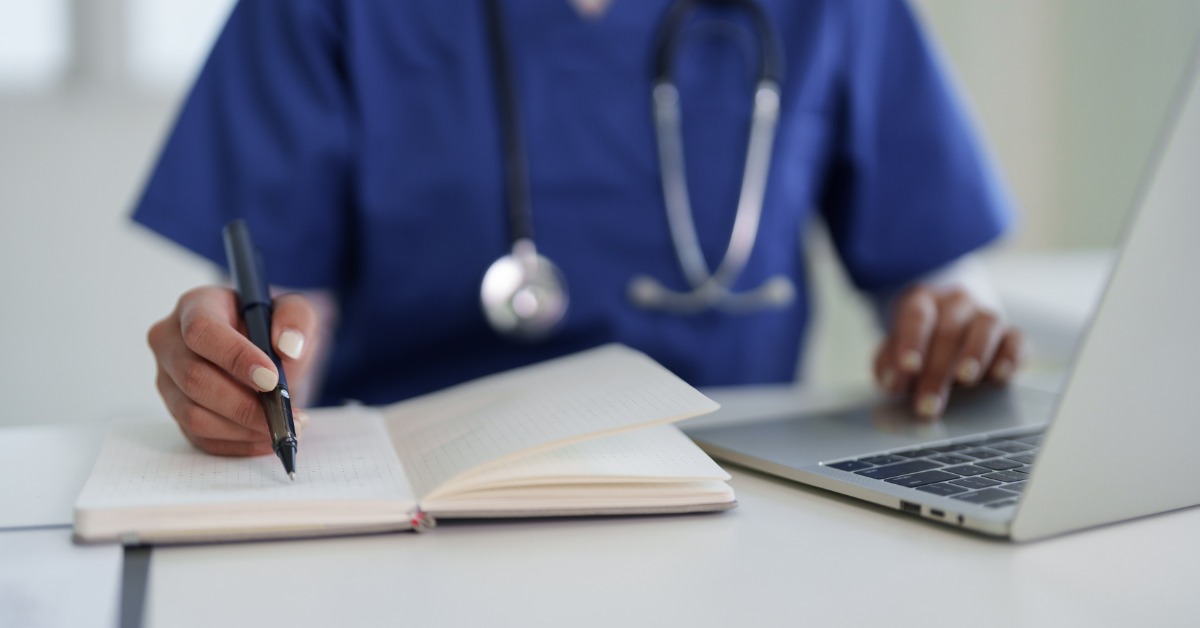 A person in blue scrubs writing in an open notebook beside a laptop, with a stethoscope around their neck.