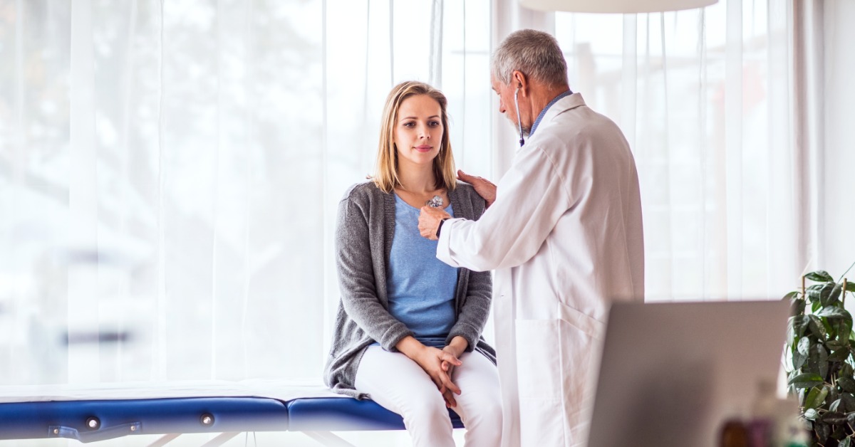 A man in a white coat using a stethoscope on a seated woman in a medical office with bright windows behind them.