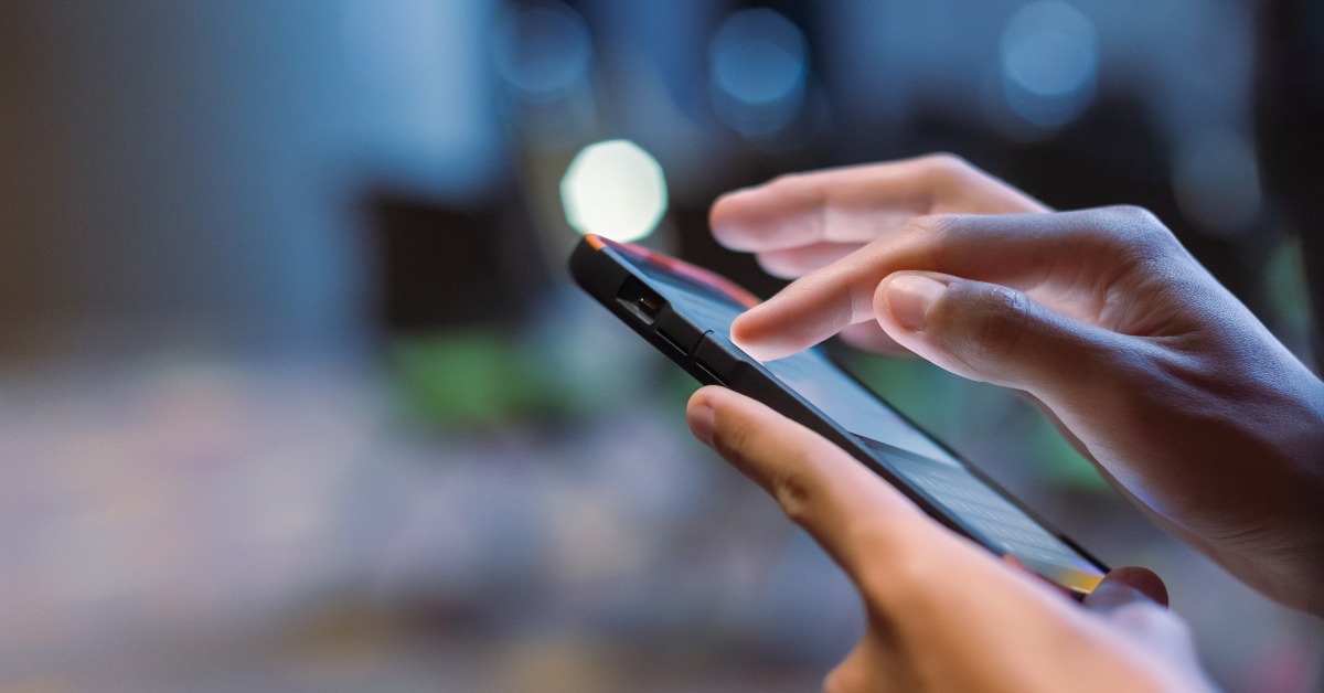 A person's hands holding and tapping a smartphone, with blurred blue lights and tables in the background.