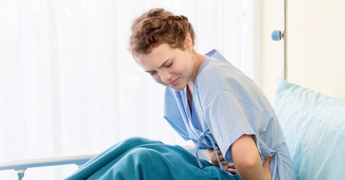 A woman wearing a blue gown sitting on a hospital bed with a blanket while holding her lower abdomen.