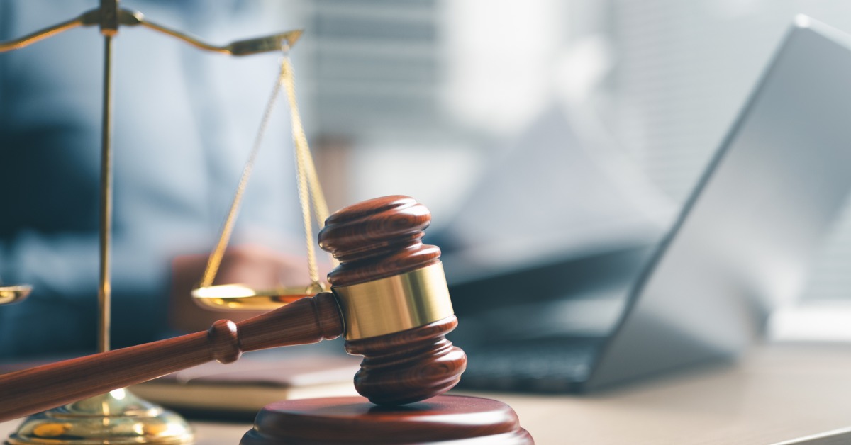 A wooden gavel beside a gold balance scale on a desk, with a laptop and blurred person in the background.