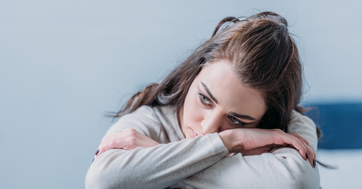 A woman resting her head on folded arms while sitting with her knees pulled up in a pale indoor room.