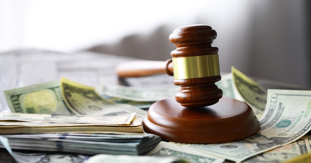 A wooden gavel resting on a round base surrounded by stacks of cash and loose bills on a table beside blurred papers.
