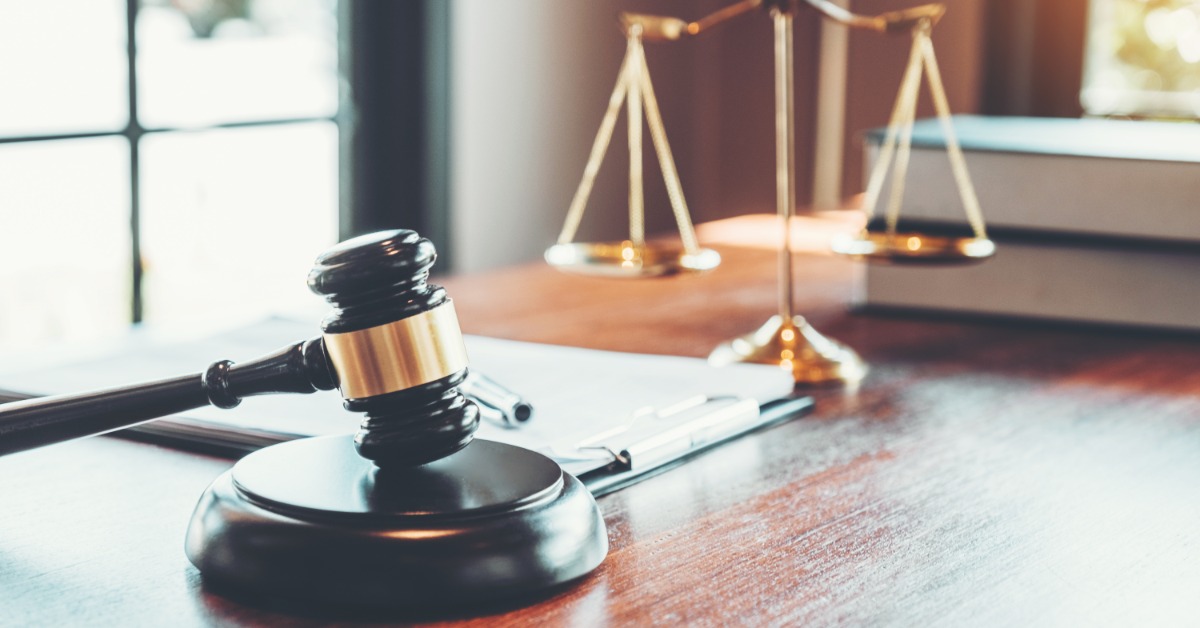 A black wooden judge's gavel resting on its base beside a clipboard, with brass balance scales and books behind it.