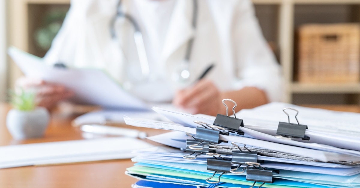 A person wearing a white coat and stethoscope writing at a desk behind a tall stack of papers held together with clips.